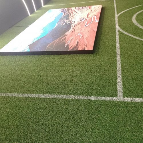 Painel de LED ligado exibindo imagem aérea de litoral, ao lado do campo de grama sintética na Casa Centauro