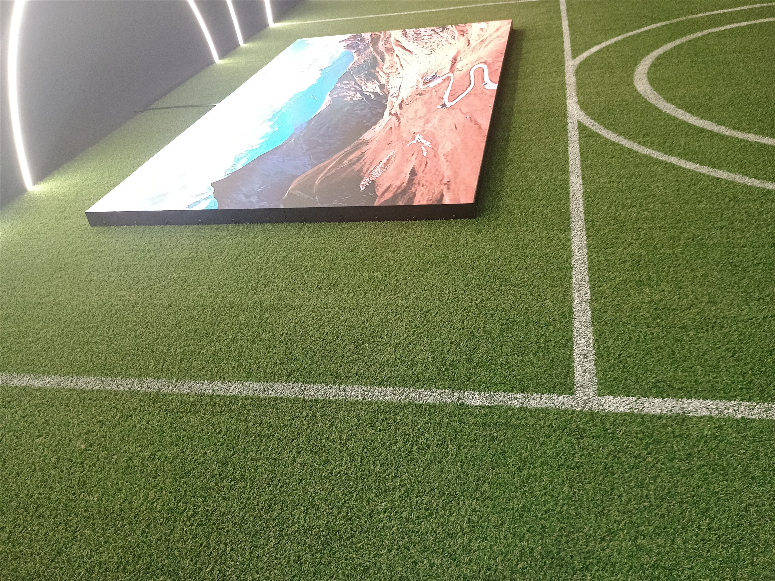 Painel de LED ligado exibindo imagem aérea de litoral, ao lado do campo de grama sintética na Casa Centauro