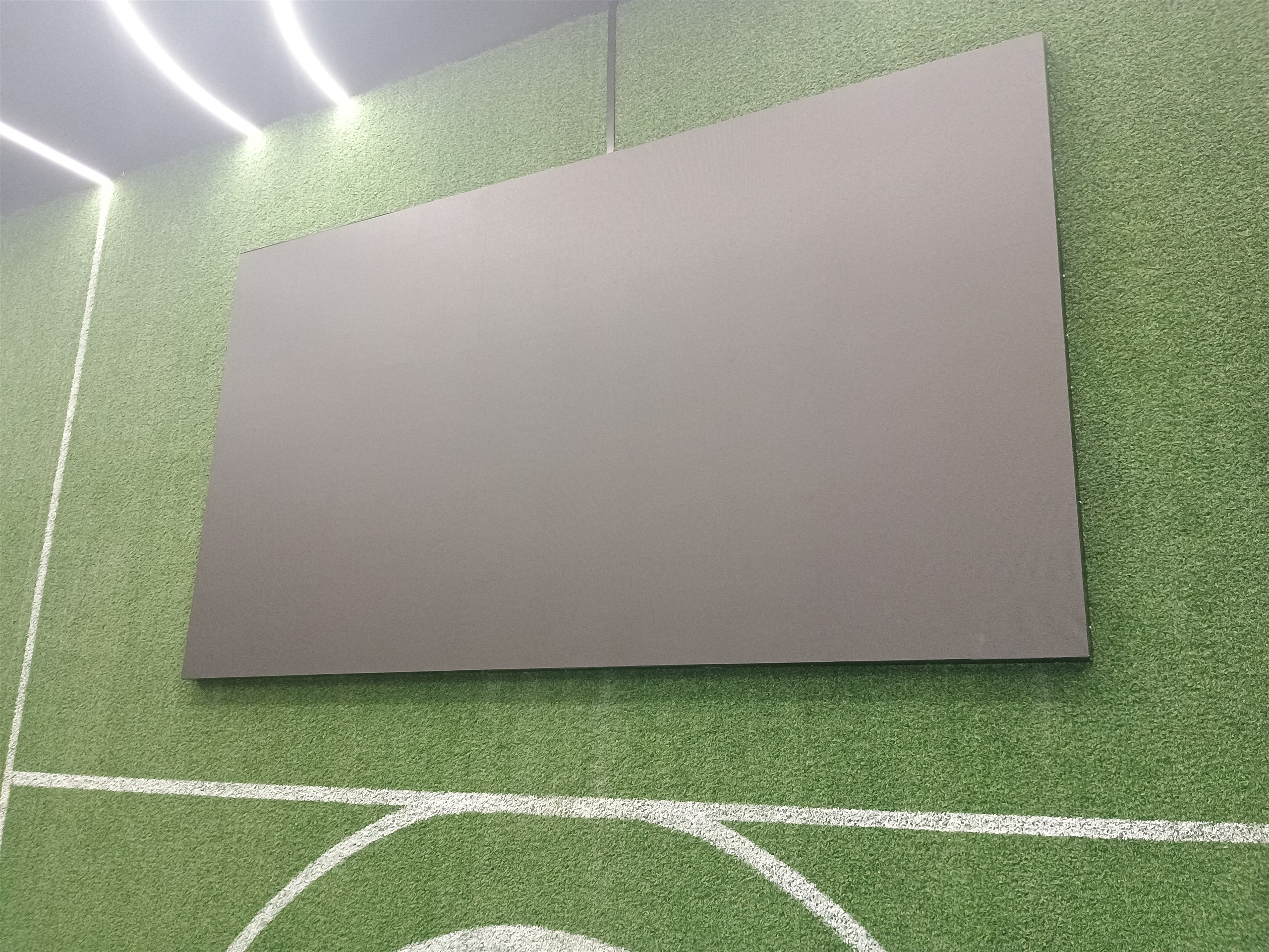 Vista frontal do painel de LED instalado em parede revestida de grama sintética com marcações de quadra