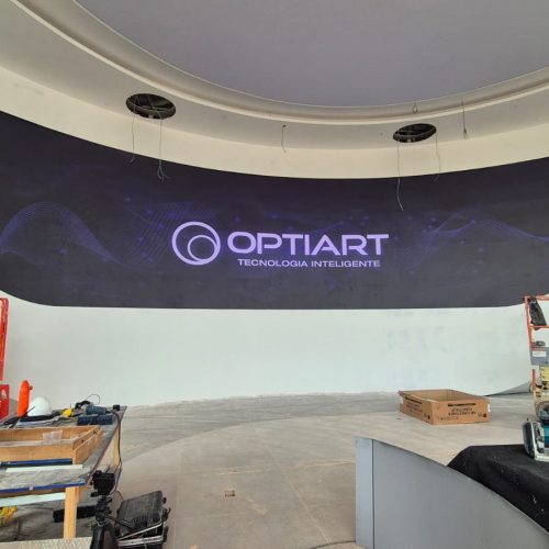 Painel de LED curvo ligado exibindo a logomarca da Optiart, embutido em parede branca no showroom da Denza em Brasília