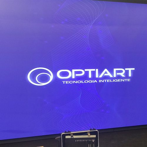 Painel de LED indoor exibindo o logotipo da Optiart na loja Yamaha Harmonia, em Brasilia (DF), ao lado de parede de madeira ripada.
