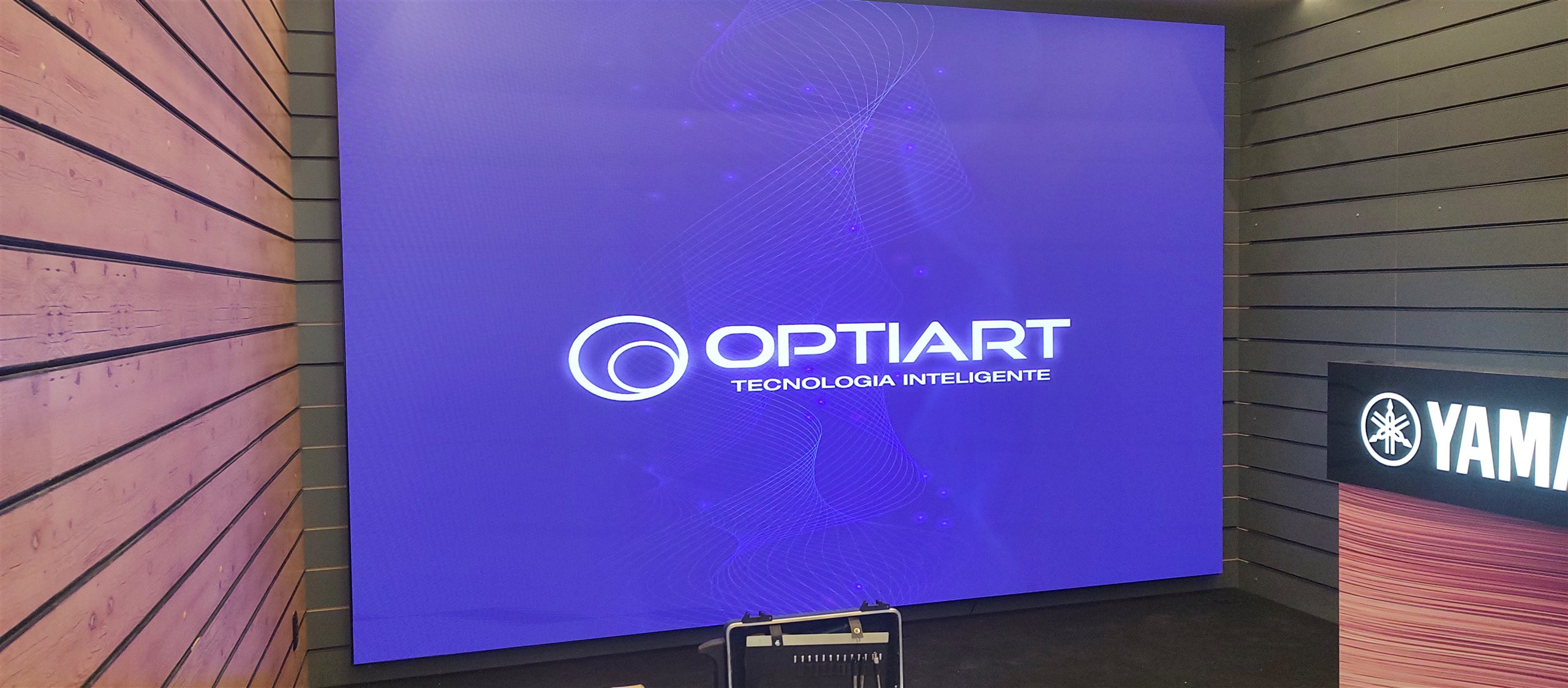 Painel de LED indoor exibindo o logotipo da Optiart na loja Yamaha Harmonia, em Brasilia (DF), ao lado de parede de madeira ripada.