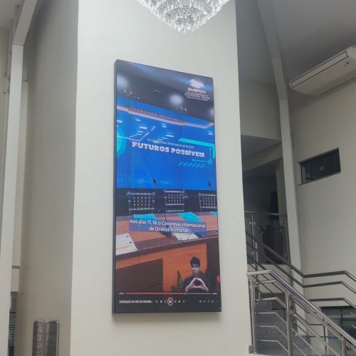 Painel de LED vertical instalado em pilar de átrio com pé-direito alto, exibindo vídeo institucional