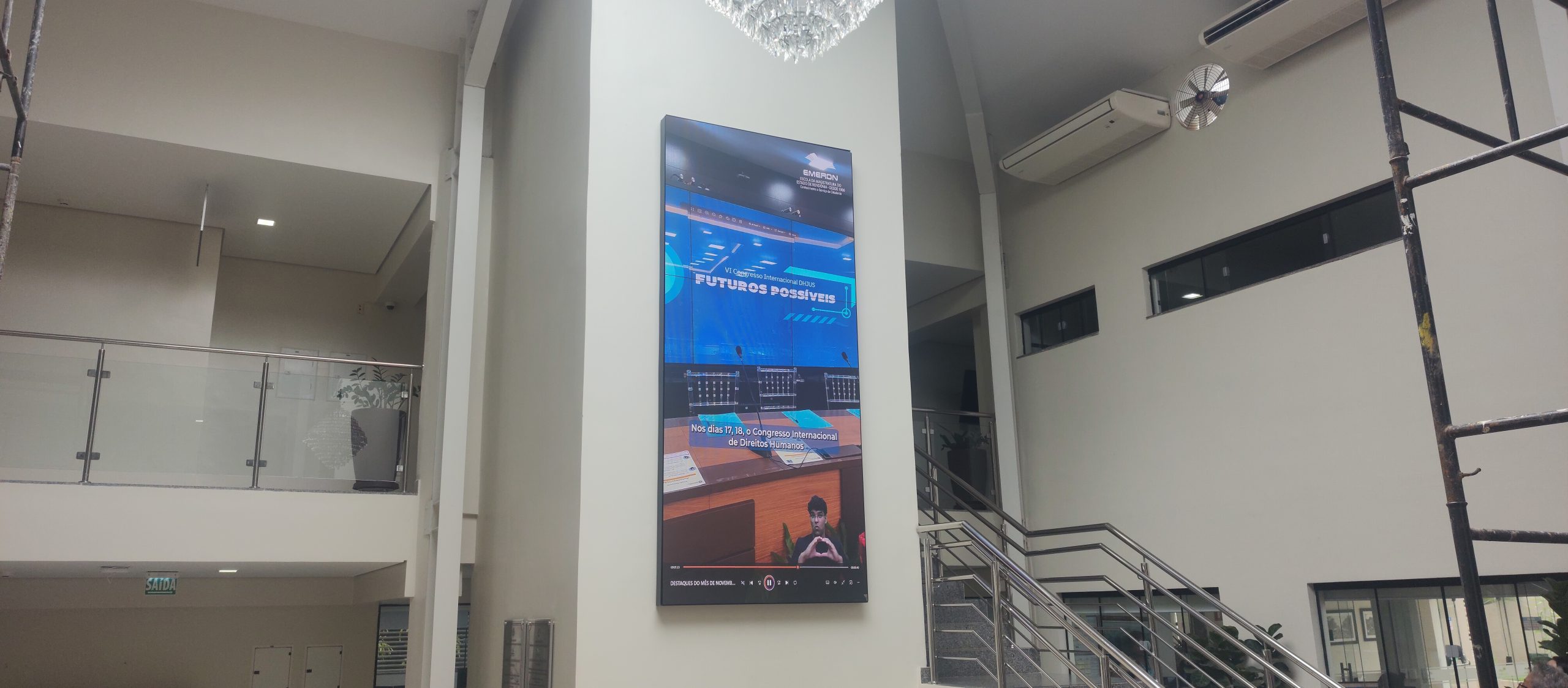 Painel de LED vertical instalado em pilar de átrio com pé-direito alto, exibindo vídeo institucional