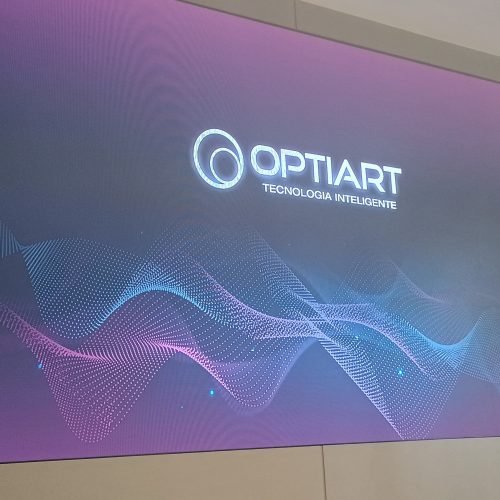 Painel de LED indoor ligado exibindo arte com o logotipo da Optiart, instalado em parede de auditório