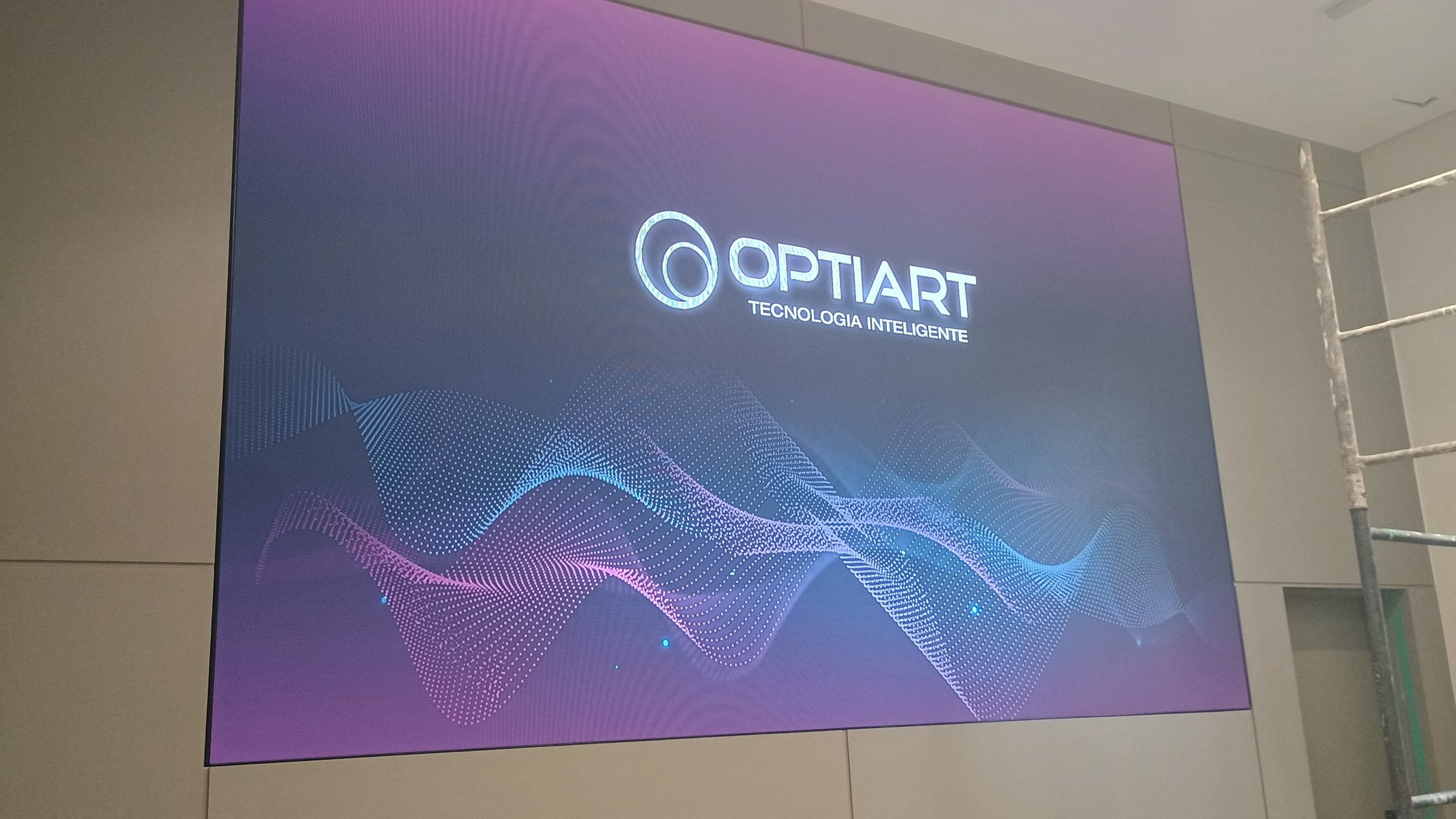 Painel de LED indoor ligado exibindo arte com o logotipo da Optiart, instalado em parede de auditório