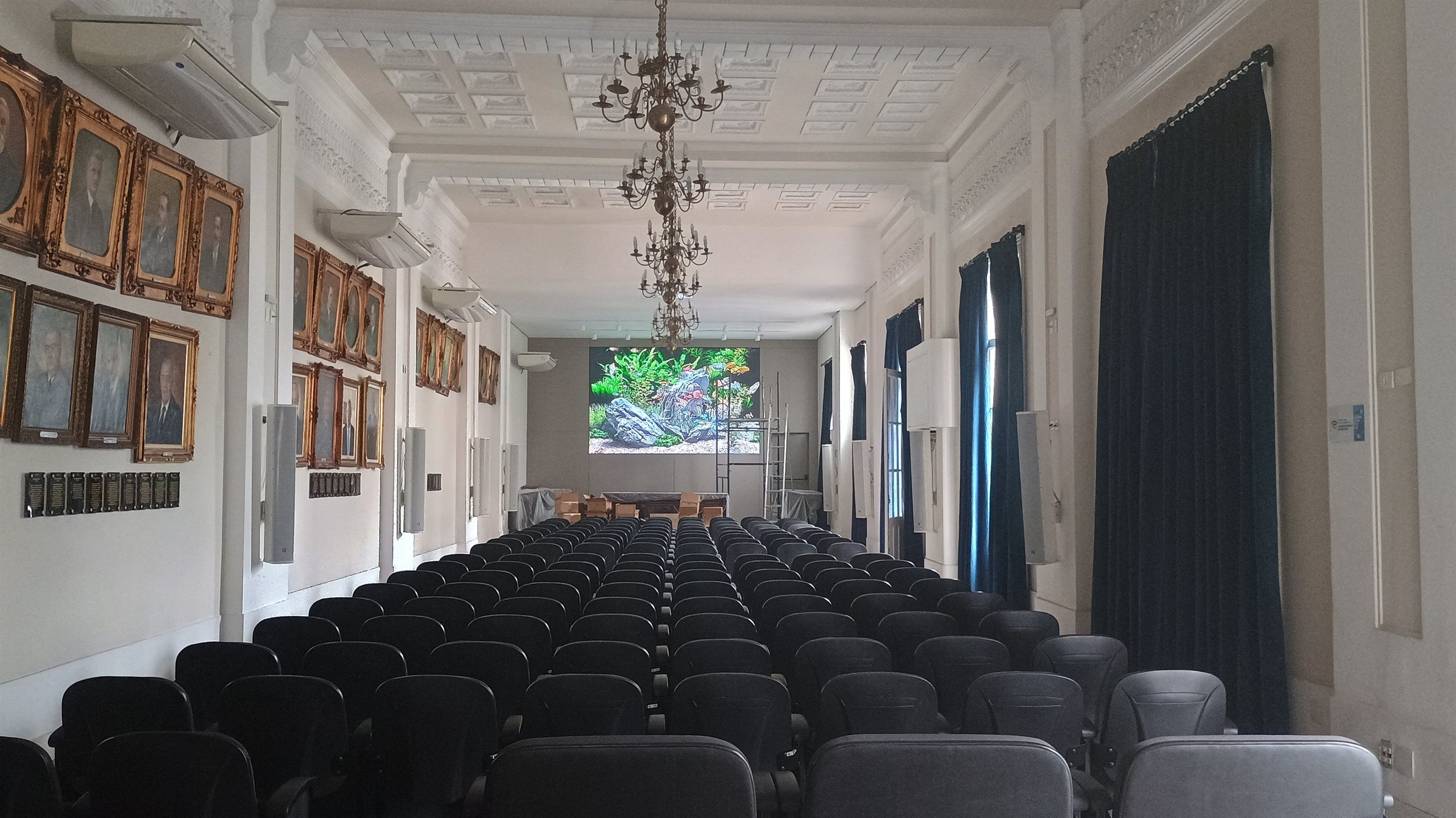 Painel de LED instalado no salão nobre da entidade empresarial de Santos, com pé-direito alto, retratos históricos emoldurados, lustre e cadeiras do auditório