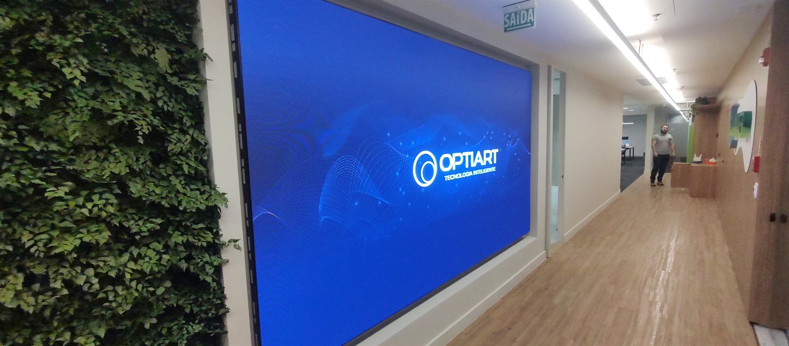 Painel de LED P2.5 em corredor corporativo exibindo conteudo institucional, parede de LED continua ao lado de jardim vertical