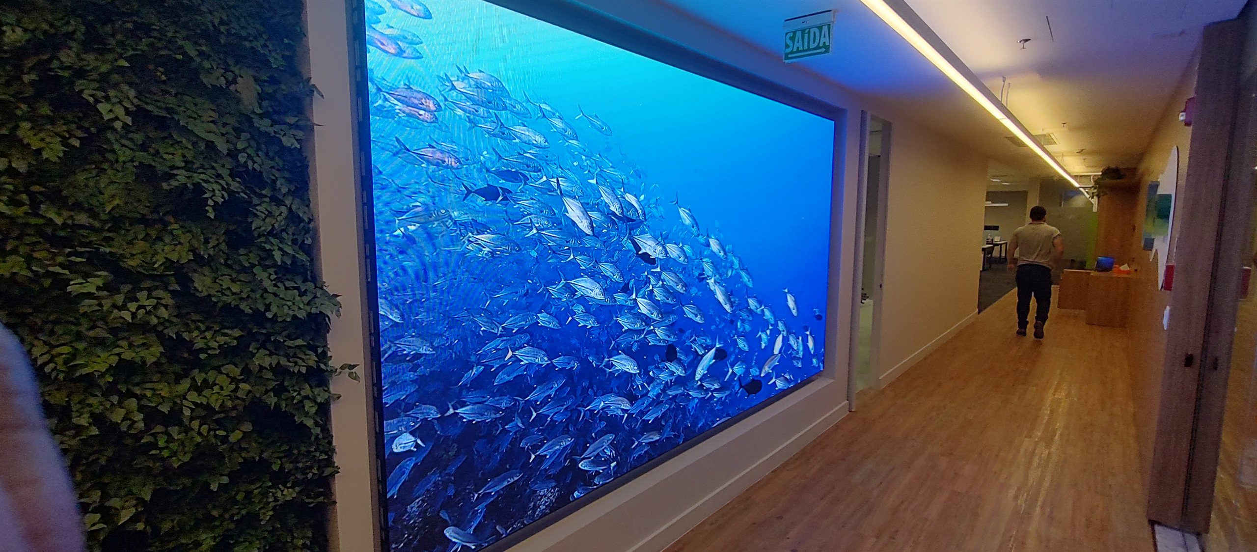 Painel de LED indoor P2.5 exibindo cena de aquario em hall corporativo, ao lado de jardim vertical