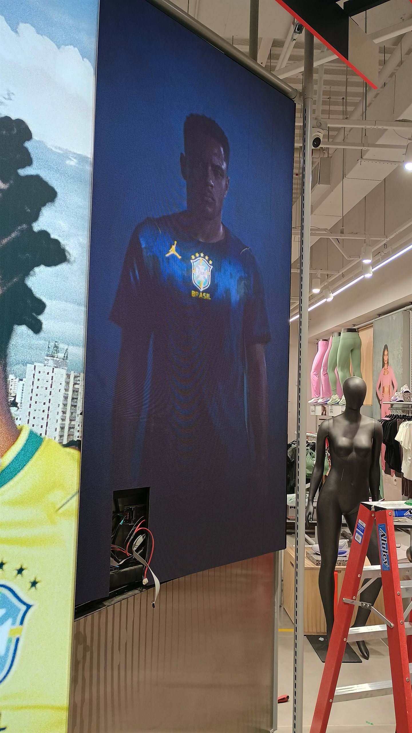 Painéis de LED verticais no piso de loja Nike exibindo atletas da seleção brasileira, integrados ao ambiente de varejo com manequins e araras de roupas