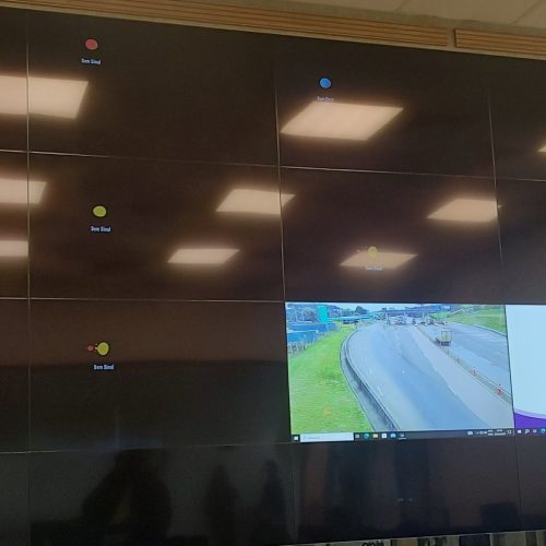 VideoWall 4x4 com dezesseis monitores instalado em nicho de painel ripado de madeira na sala de controle de uma concessionária de rodovias, com janela de conteúdo aberta sobre a matriz