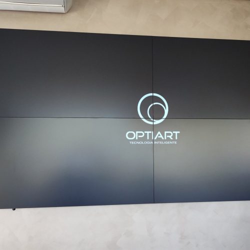 VideoWall 2x2 com quatro monitores de 55 polegadas ligado com a marca Optiart na tela, instalado em parede de cimento queimado do stand de vendas