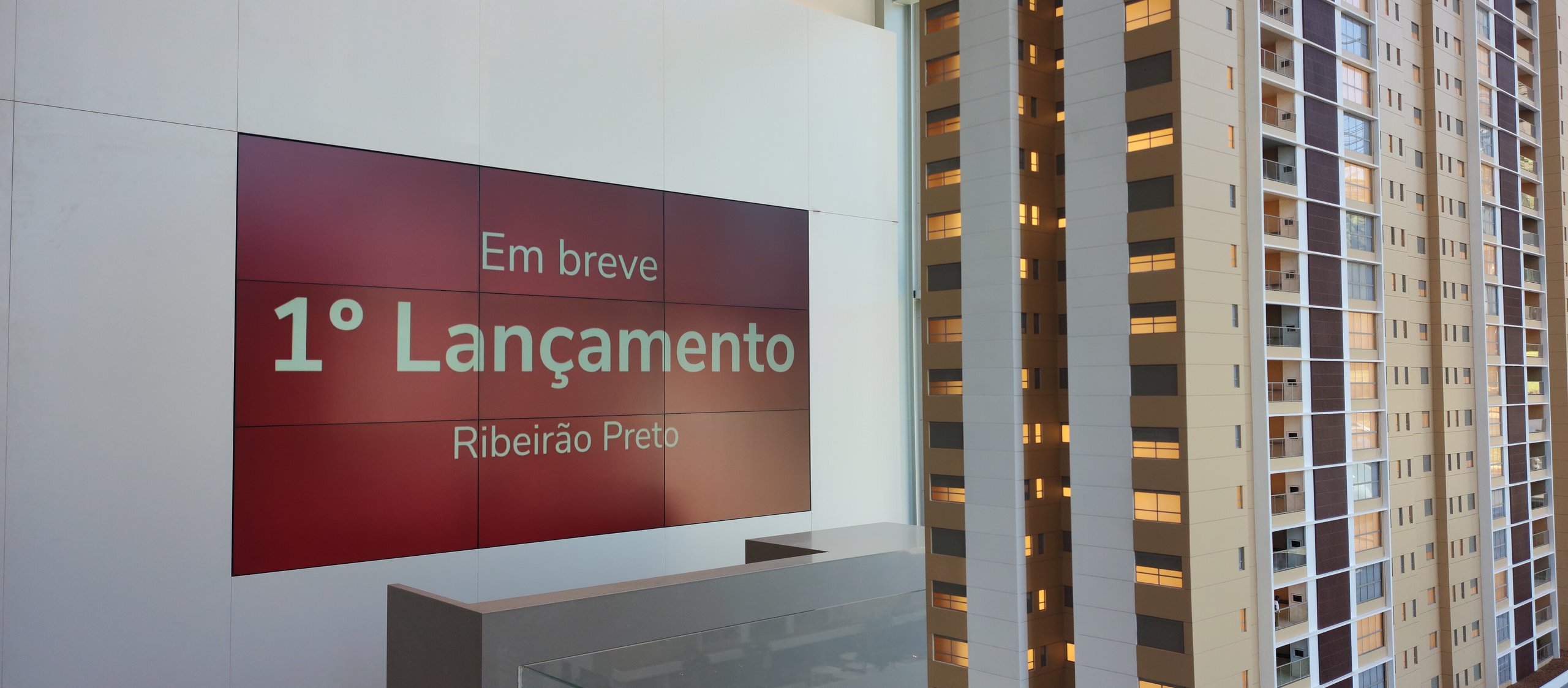 VideoWall 3x3 exibindo peça de lançamento imobiliário no hall do stand de vendas, com as torres do empreendimento ao fundo