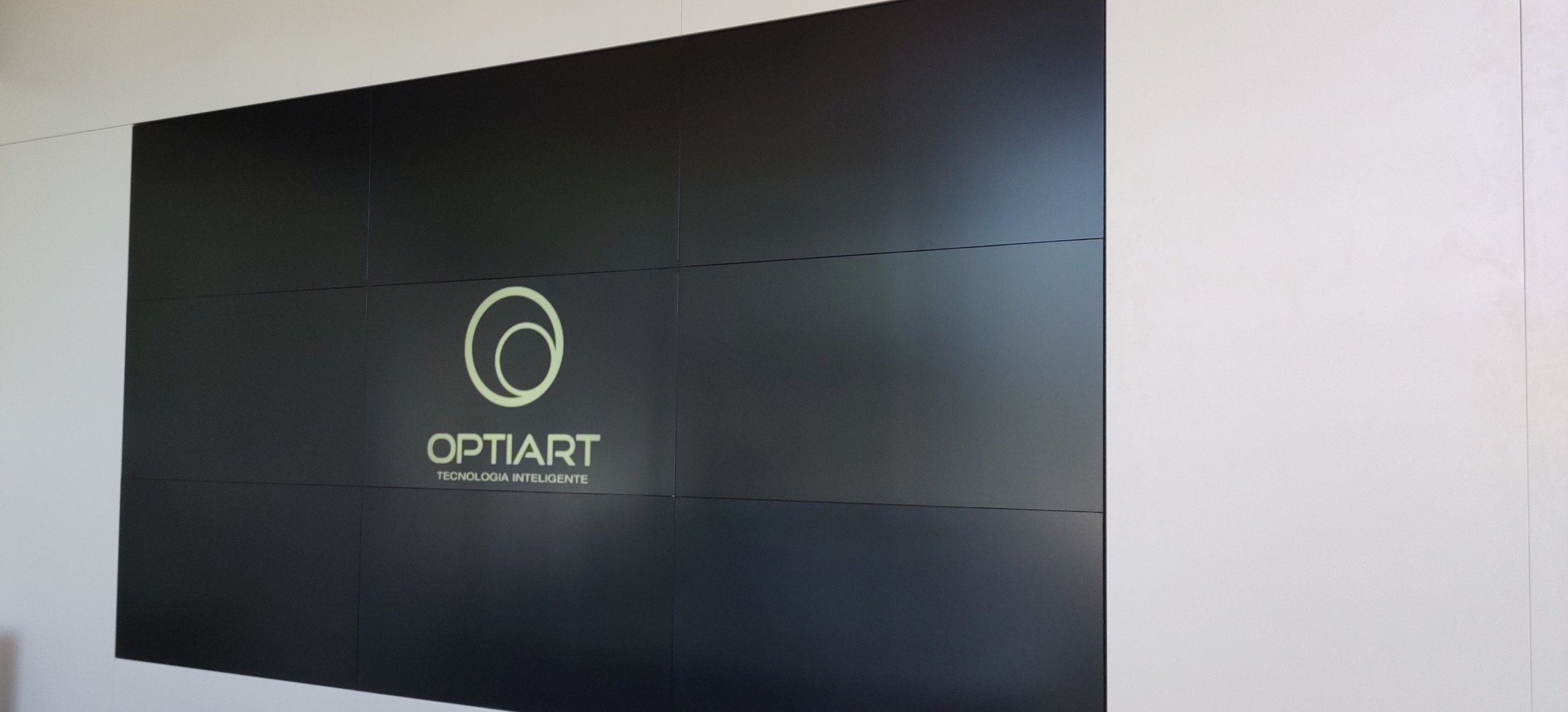 Segundo videowall 3x3 do stand de vendas ligado com a marca Optiart na tela, instalado sobre painel de fechamento branco