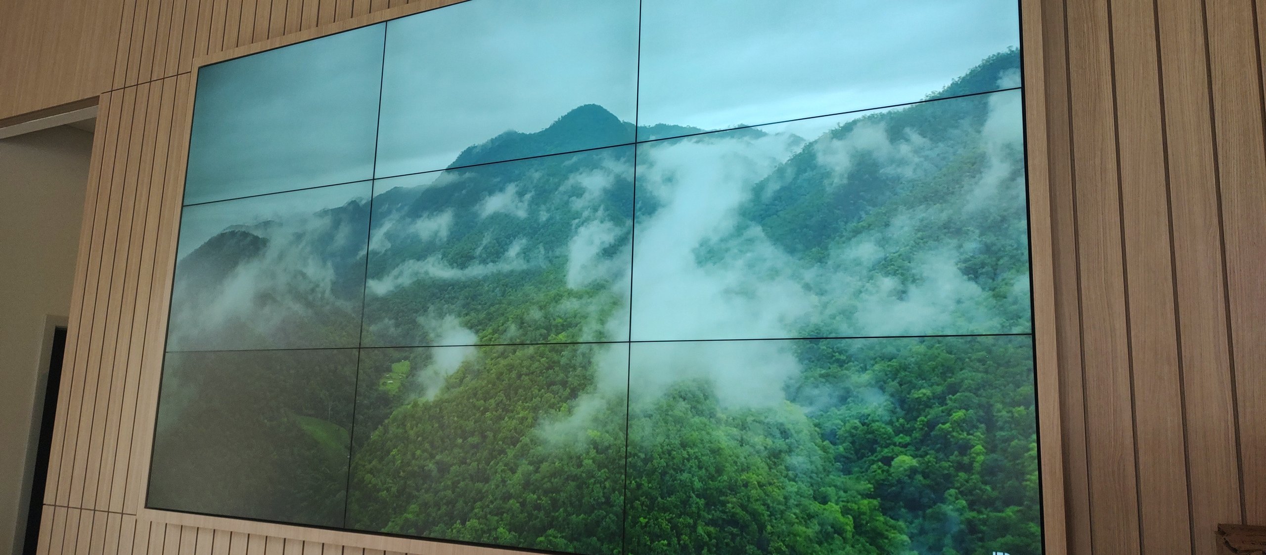 VideoWall 3x3 com nove monitores de 55 polegadas exibindo imagem de natureza em tela cheia, embutido em nicho de painel ripado de madeira no stand de vendas