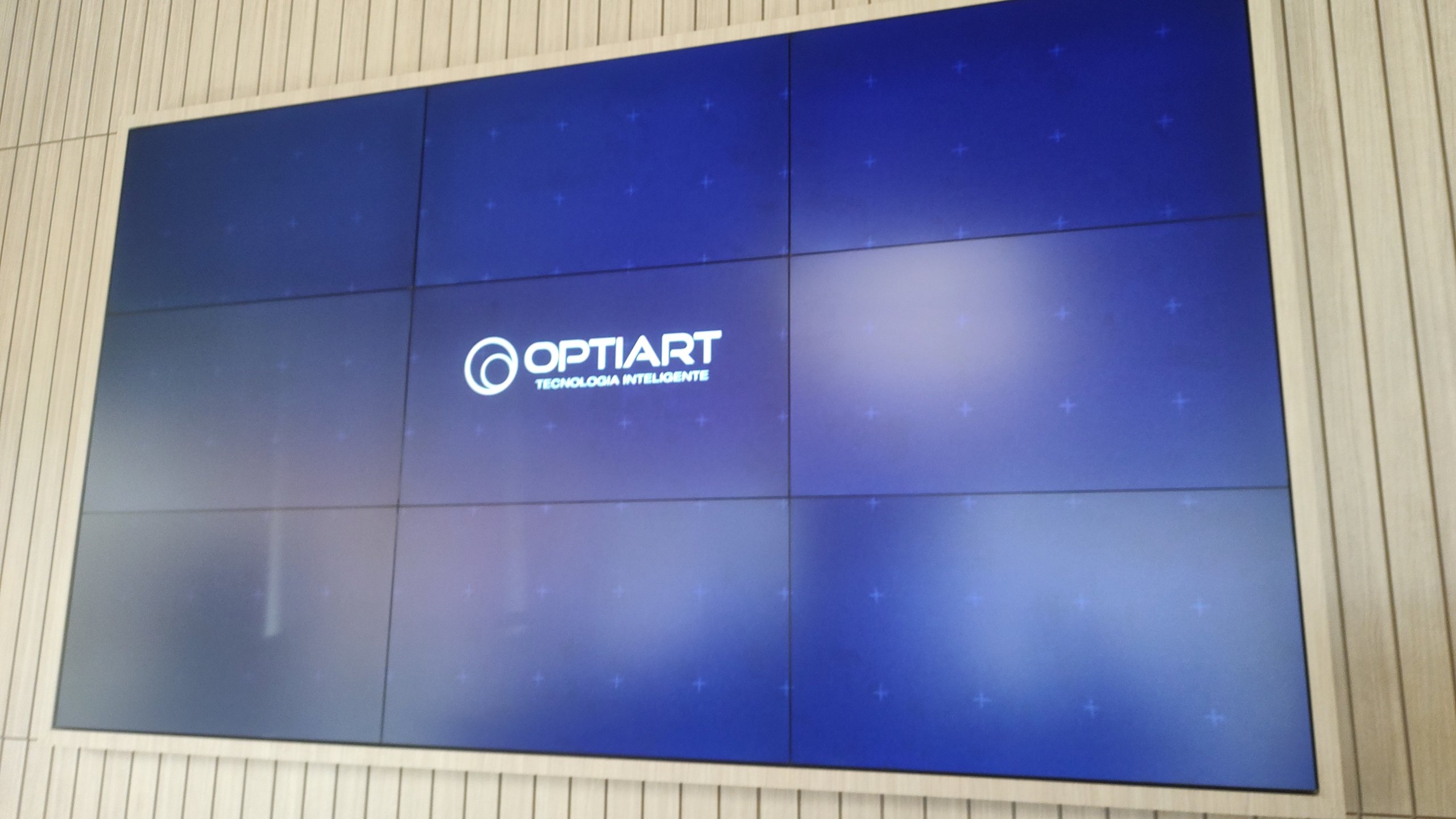 VideoWall 3x3 ligado com a marca Optiart na tela, instalado em nicho de painel ripado de madeira