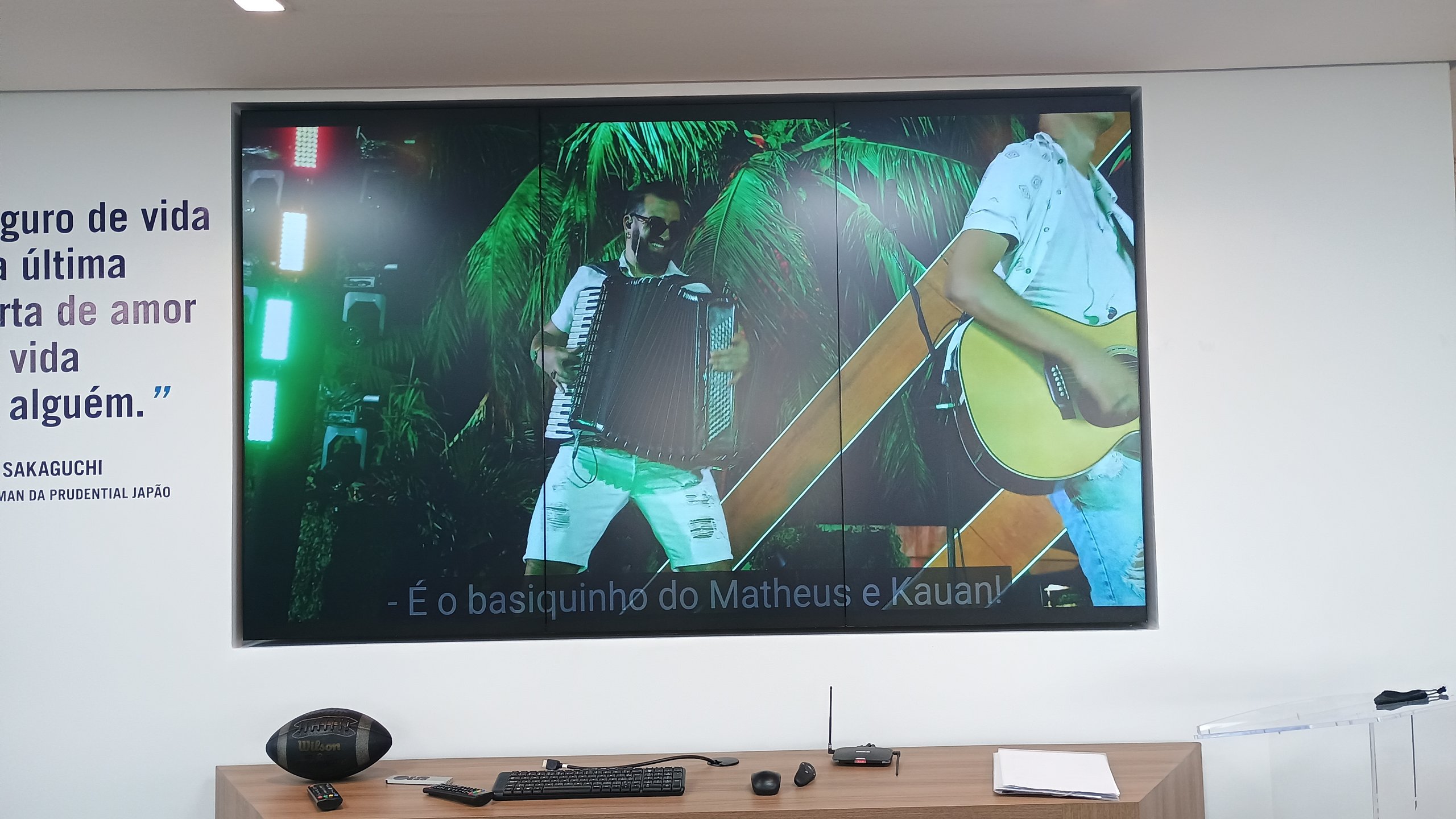 VideoWall 1x3 com três monitores verticais embutidos em nicho exibindo vídeo em tela cheia na recepção da Prudential em Goiânia, com frase institucional na parede ao lado