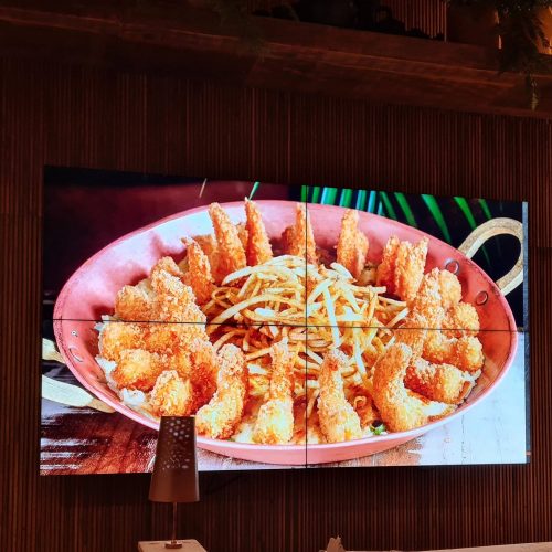 VideoWall 2x2 ligado no salão de um restaurante, exibindo a foto de um prato em tela cheia sobre parede ripada de madeira