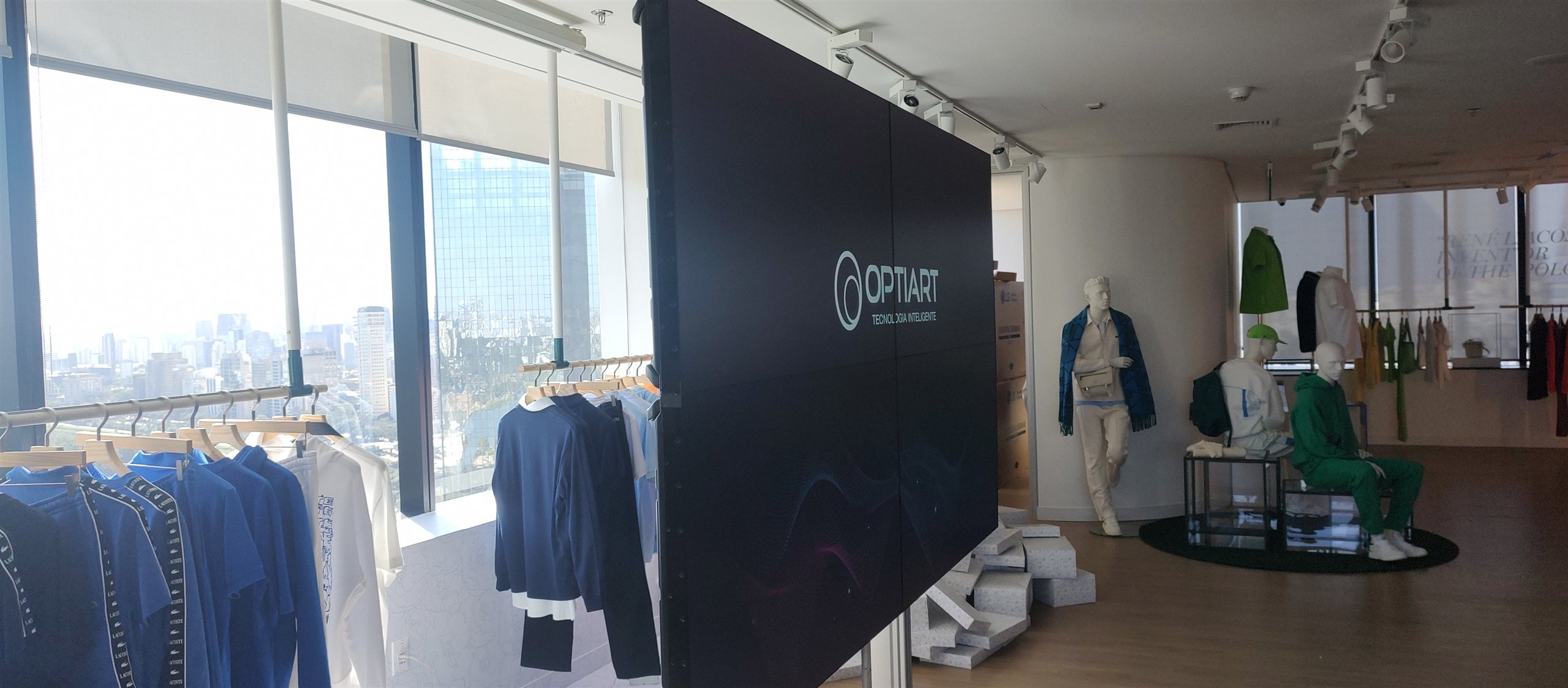 VideoWall sobre pedestal com rodízios integrado ao ambiente do showroom da Lacoste, com araras de roupas e manequins
