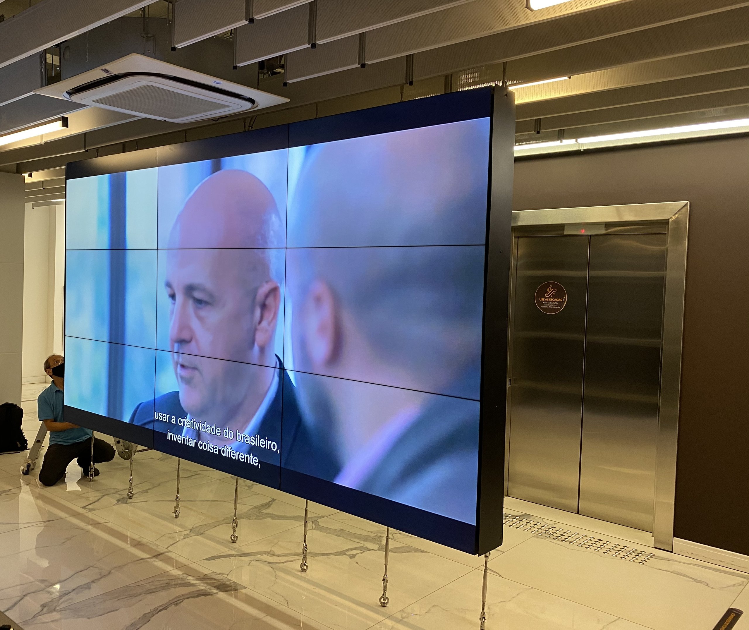 VideoWall 3x3 montado em estrutura autoportante junto aos elevadores, com tirantes de fixação no piso