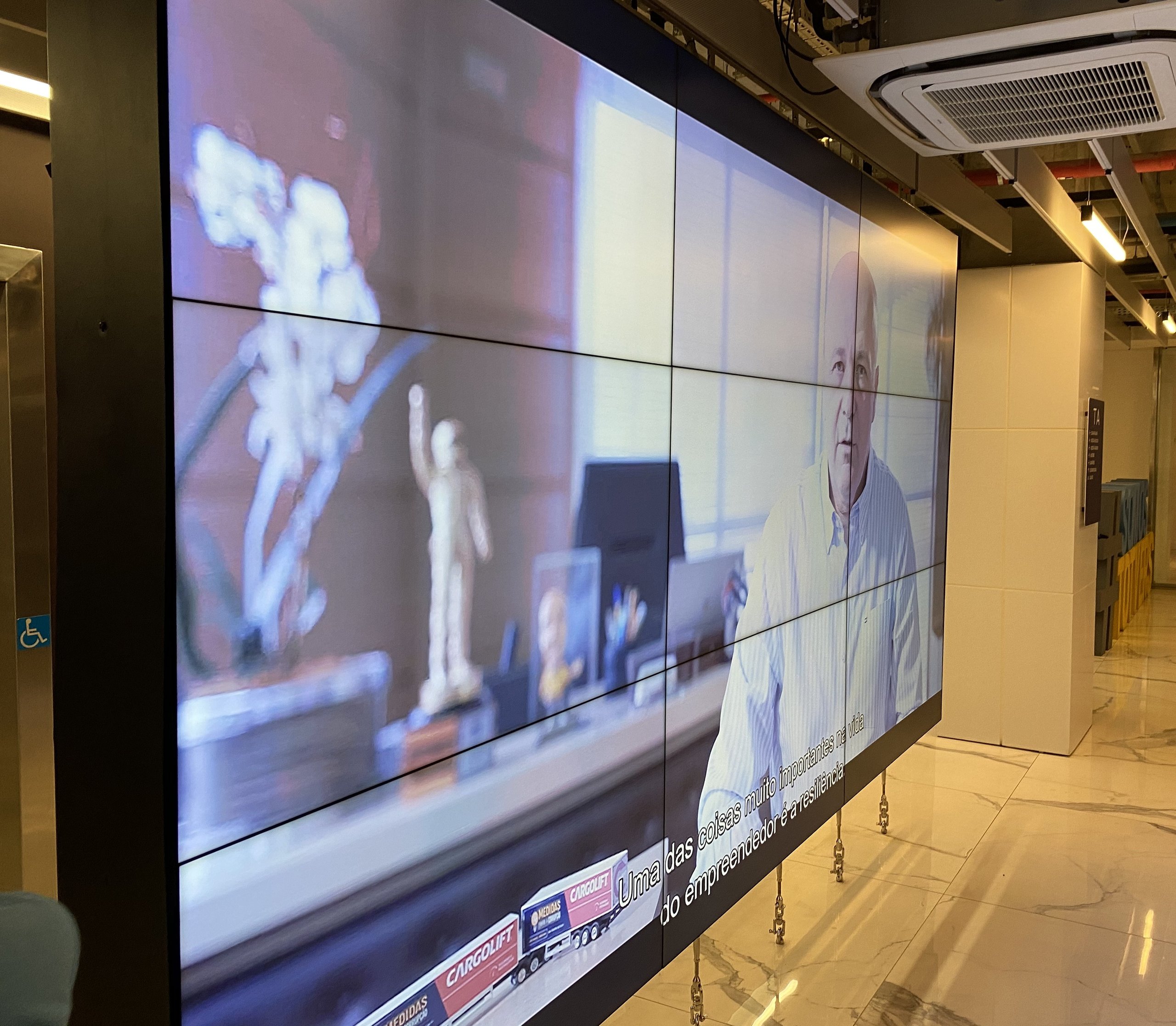 Vista em perspectiva do VideoWall 3x3 no corredor de circulação corporativa, com piso em mármore e telas ligadas