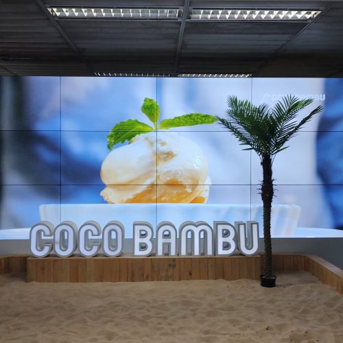 VideoWall 3x4 do Coco Bambu no Aeroporto de Guarulhos exibindo imagem de sobremesa, com letreiro Coco Bambu à frente