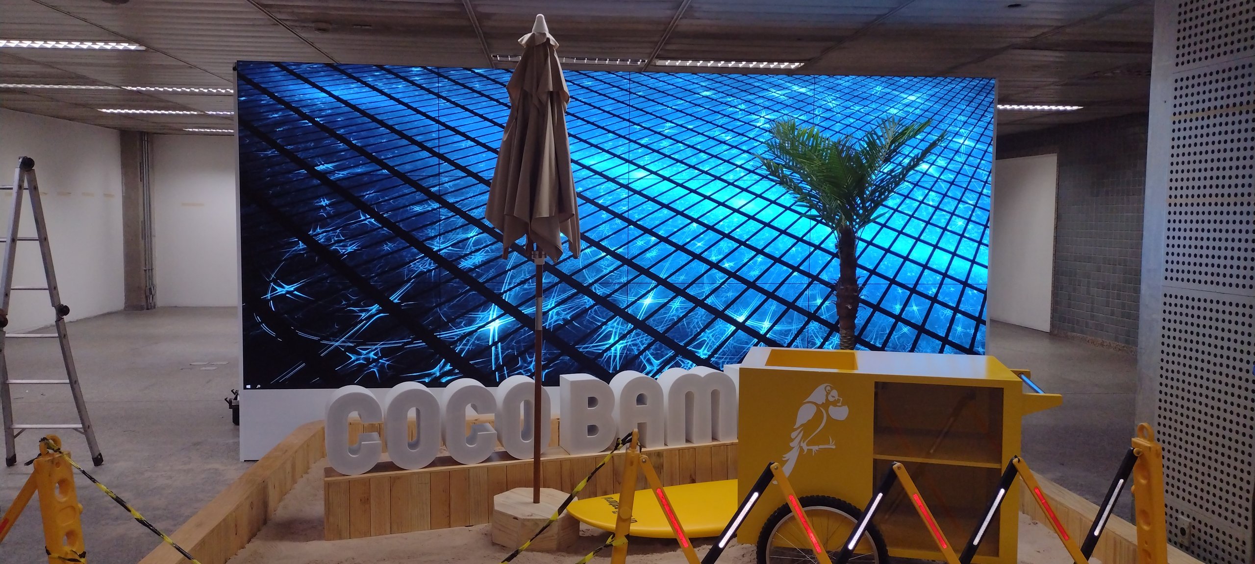 Vista frontal do VideoWall 3x4 móvel com conteúdo em azul, letreiro Coco Bambu e ambientação de praia no estande